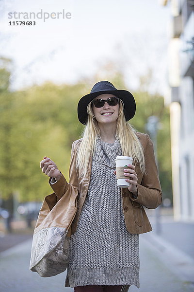 Hübsche blonde Frau mit Hut  Sonnenbrille und Kaffee zum Spazierengehen im Stadtzentrum.