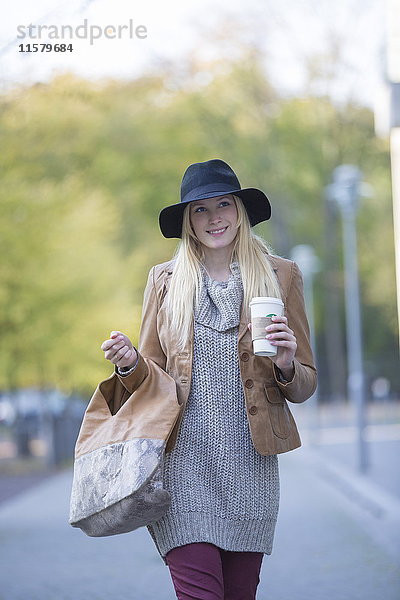 Hübsche blonde Frau mit Hut und Kaffee für einen Spaziergang im Stadtzentrum.