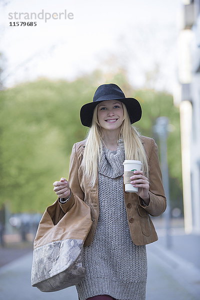 Hübsche blonde Frau mit Hut und Kaffee  die vor der Kamera lächelt und im Stadtzentrum spazieren geht.