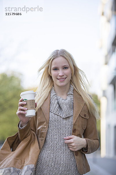 Hübsche blonde Frau mit Café zum Spazierengehen im Stadtzentrum und lächelndem Blick auf die Kamera.