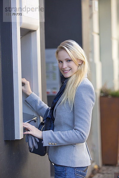 Hübsche blonde Frau  die einen Geldautomaten benutzt und die Kamera anlächelt.