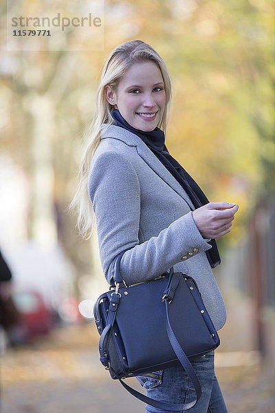 Hübsche elegante Frau geht mit Handtasche im Stadtzentrum und lächelt in die Kamera.