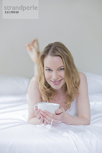Hübsche blonde Frau mit einer Tasse Tee im Bett  lächelnd vor der Kamera.