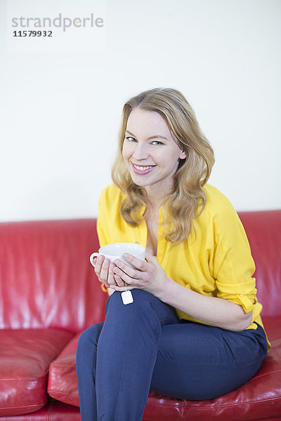 Porträt einer schönen blonden Frau  die eine Tasse Tee auf dem Sofa genießt und vor der Kamera lächelt.
