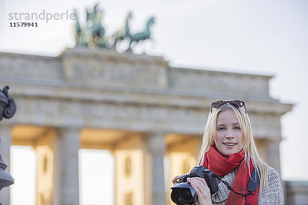 Hübsche blonde Frau fotografiert vor dem Brandenburger Tor in Berlin und schaut in die Kamera.