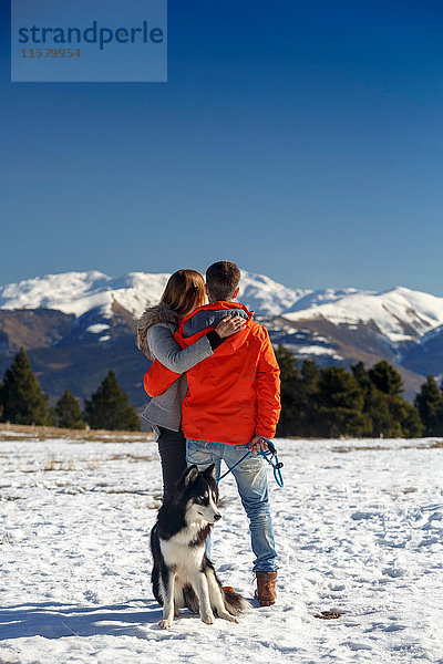 Rückansicht eines romantischen Paares mit Hund beim Blick auf die schneebedeckte Berglandschaft