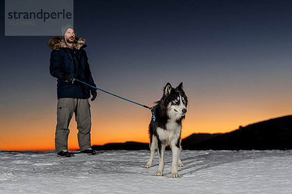 Erwachsener Mann steht nachts mit Hund im Schnee