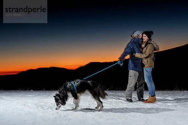 Romantisches Paar mit Hund bei Nacht im Schnee