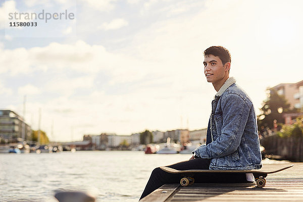 Junger Mann sitzt am Fluss  Skateboard neben ihm  Bristol  UK