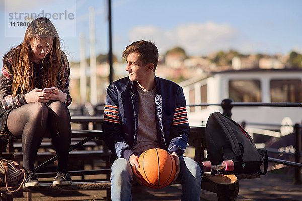 Zwei Freunde sitzen im Freien  junger Mann hält Basketball  junge Frau benutzt Smartphone  Bristol  UK