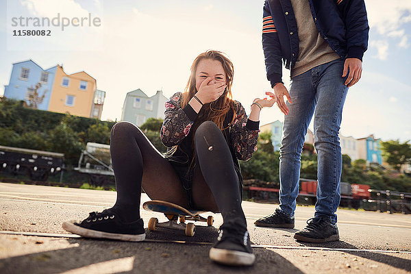 Zwei Freunde albern im Freien herum  junge Frau sitzt auf einem Skateboard  lachend  niedrige Sektion  Bristol  UK
