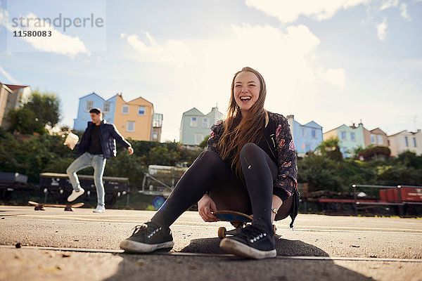 Zwei Freunde albern im Freien herum  junge Frau sitzt auf einem Skateboard und lacht  Bristol  UK