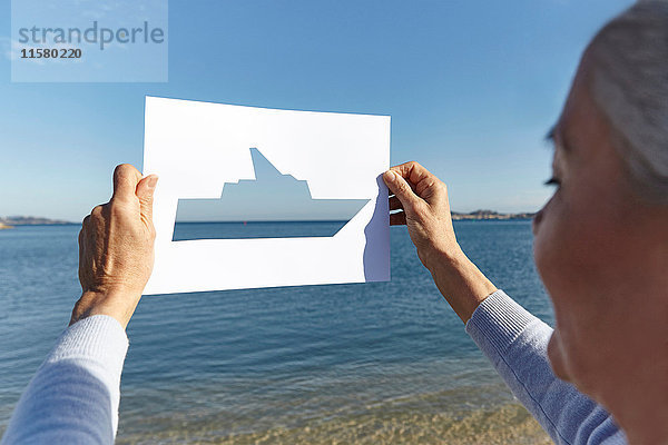 Ältere Frau steht mit dem Gesicht zum Meer und hält ein Papier mit einem Ausschnitt aus einem Boot