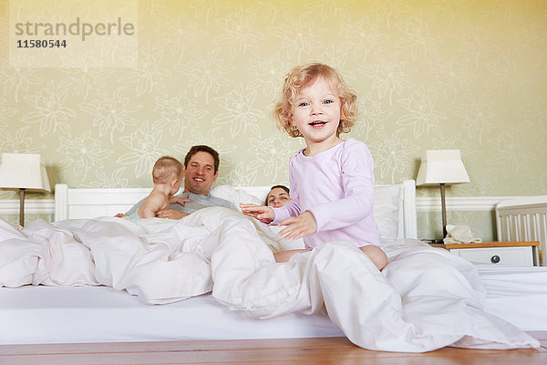 Porträt eines süßen weiblichen Kleinkindes  das mit Eltern und kleiner Schwester auf dem Bett sitzt