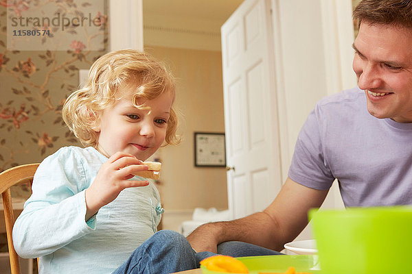 Mittelgroßer erwachsener Mann und Kleinkind-Tochter essen Brot am Küchentisch
