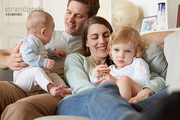 Mittlere erwachsene Eltern kauerten mit Baby und Kleinkind-Tochter auf dem Sofa