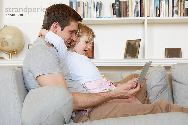 Weibliches Kleinkind sitzt mit seinem Vater auf dem Sofa und schaut auf ein digitales Tablett