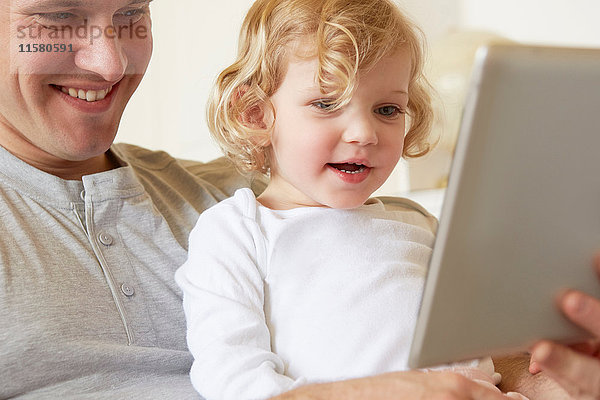 Weibliches Kleinkind sitzt auf dem Knie des Vaters und benutzt digitales Tablett