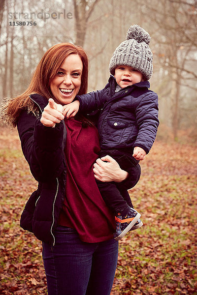Mittelgroße erwachsene Frau  die einen Kleinkind-Sohn trägt und in den Herbstwald zeigt