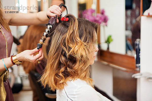 Friseur mit Lockenstab am langen braunen Haar des Kunden im Salon