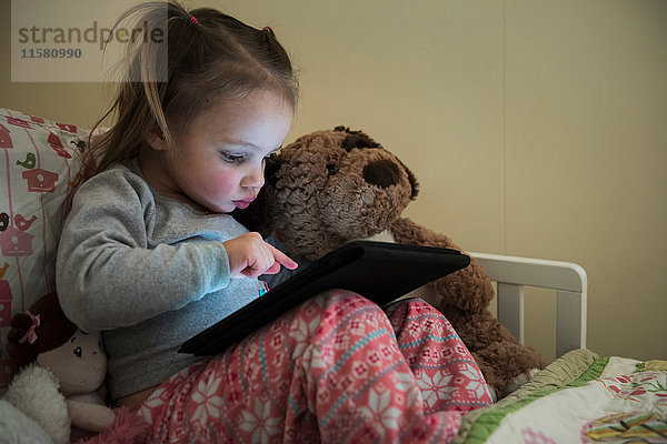 Weibliches Kleinkind sitzt aufrecht im Bett und benutzt Touchscreen auf digitalem Tablett