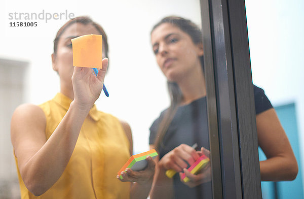 Zwei Geschäftsfrauen kleben Klebezettel an Büro-Glaswand