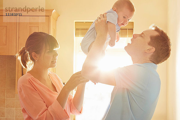 Reifer Mann mit Frau hält kleinen Sohn in sonnenbeschienener Küche hoch