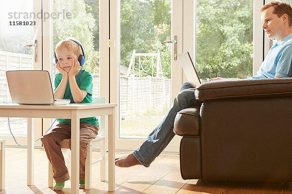 Junge am Schreibtisch und Vater auf dem Sofa mit Laptops