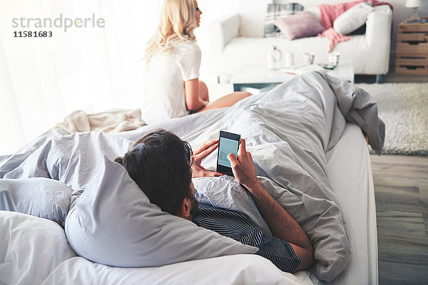Mann im Bett mit Smartphone  Frau am Ende des Bettes sitzend