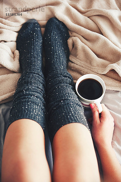 Frau sitzt auf dem Bett  trägt warme Socken  hält eine Tasse Kaffee  niedriger Schnitt  Draufsicht