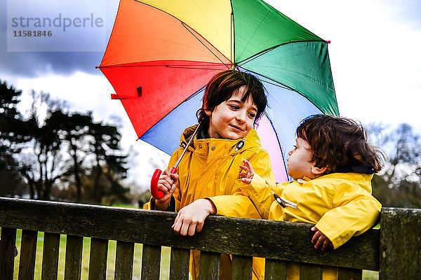 Draufsicht auf den kleinen Jungen und Bruder in gelben Anoraks mit Regenschirm auf der Parkbank