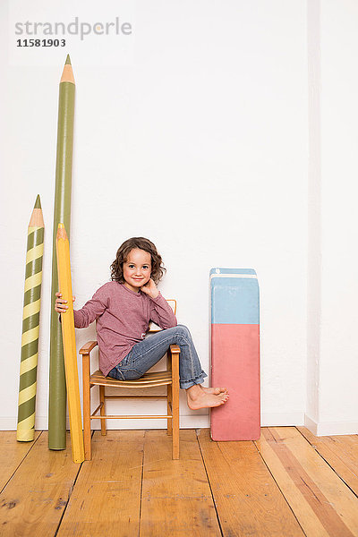 Junges Mädchen sitzt auf einem Stuhl  hält einen Bleistift in Riesengröße in der Hand  neben sich an der Wand lehnt ein riesengroßes Briefpapier