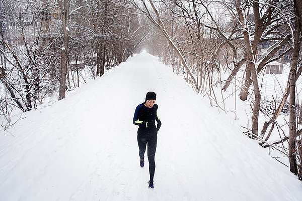 Junge Läuferin mit Strickmütze läuft in einem von Bäumen gesäumten  schneebedeckten Park