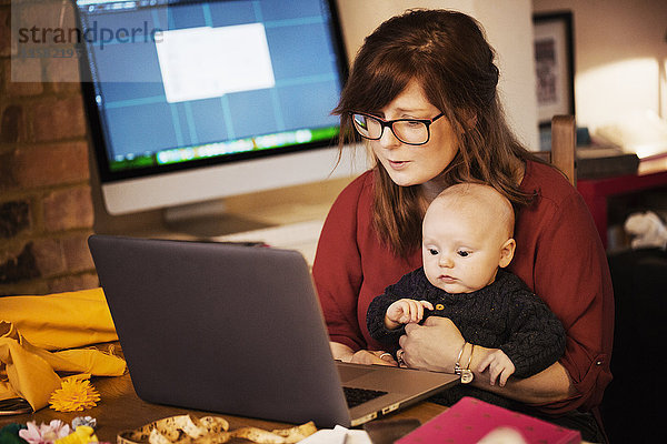 Eine Frau sitzt mit einem Baby auf dem Schoß  das einen Computer-Laptop benutzt  wobei beide Personen aufmerksam auf den Bildschirm schauen.
