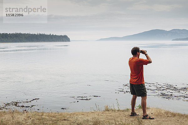 Mann steht am Strand und schaut durch ein Fernglas  San Juan Inseln in der Ferne  Washington  USA.