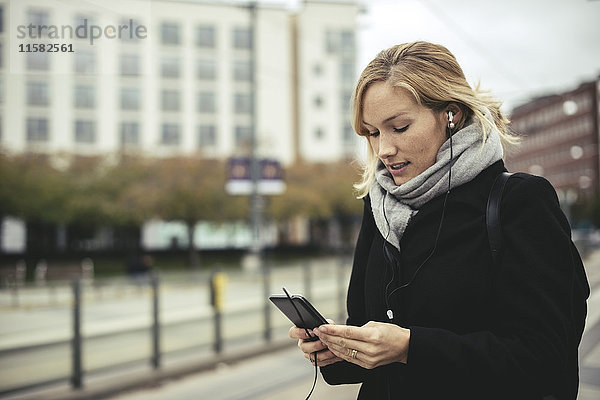 Mittlere erwachsene Geschäftsfrau beim Musikhören über Smartphone an der Straßenbahnstation