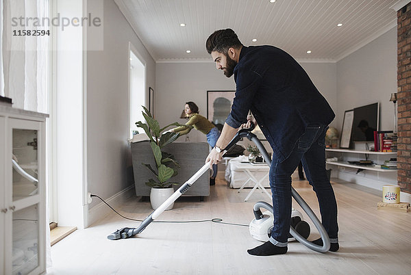 Mann saugt Boden  während Frau im Wohnzimmer zu Hause arbeitet