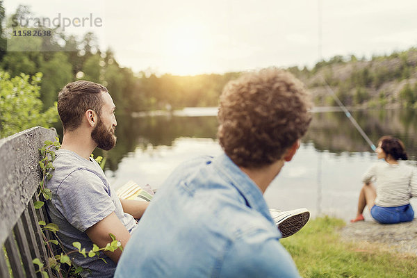 Männer sitzen auf einer Holzbank  während eine Freundin am Seeufer fischt.