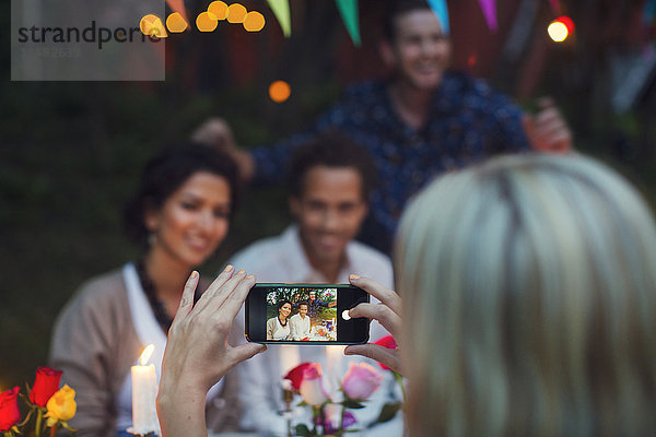 Abgeschnittenes Bild einer Frau  die glückliche Freunde durch ein Smartphone auf einer Gartenparty fotografiert.