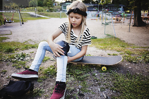 Mädchen  das auf dem Skateboard im Park sitzt und Kniepolster bindet.