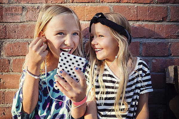 Fröhliche Mädchen  die Musik über das Smartphone hören  während sie an der Wand sitzen.