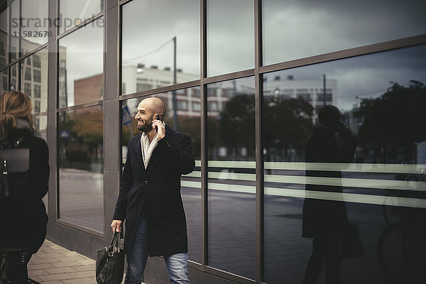 Lächelnder Geschäftsmann im Gespräch mit dem Handy beim Spaziergang durch das Stadtgebäude