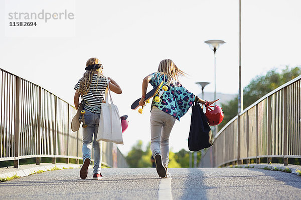 Volle Länge von Freunden  die Skateboards halten  während sie auf der Brücke gegen den klaren Himmel laufen.