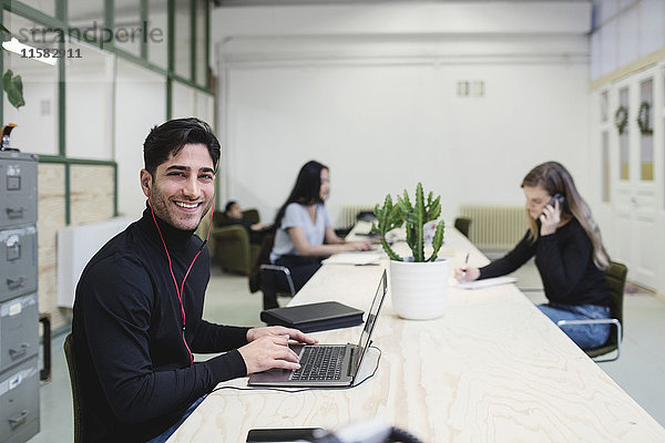 Porträt eines glücklichen jungen Mannes am Schreibtisch mit Kollegen im Hintergrund