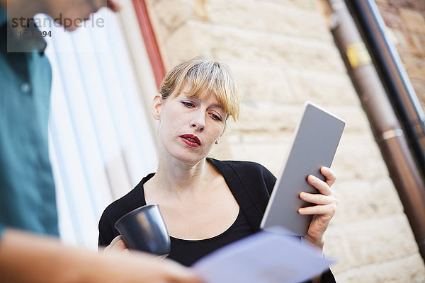 Mittlere erwachsene Geschäftsfrau mit digitalem Tablett und Kaffeetasse im Gespräch mit Kollegen außerhalb des Büros