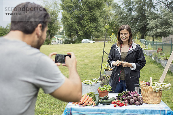 Mittlerer Erwachsener Mann  der eine Frau fotografiert  die an einem Tisch voller Gartengemüse steht.