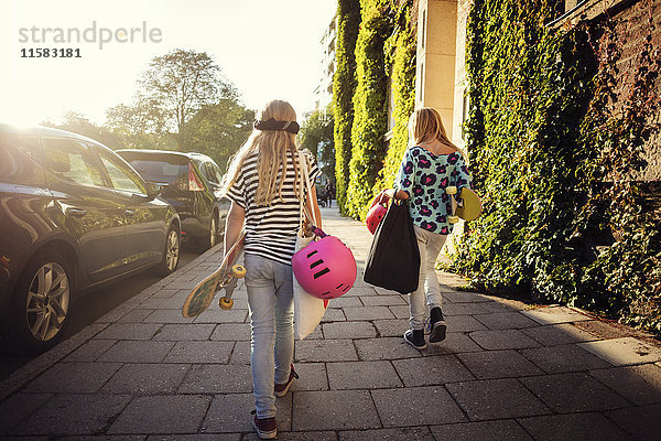 Rückansicht von Mädchen mit Skateboards  die an sonnigen Tagen mit geparkten Autos auf dem Bürgersteig laufen.