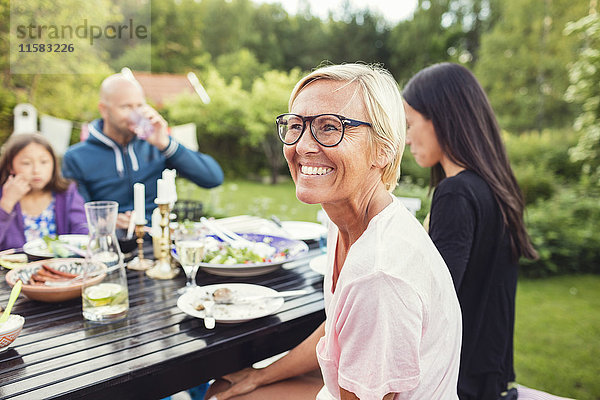 Glückliche Frau sitzt mit Freunden und Familie am Esstisch im Garten während der Gartenparty.