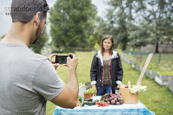 Mittlerer Erwachsener Mann  der eine Frau fotografiert  die im Stadtgarten neben frisch geerntetem Gemüse steht.