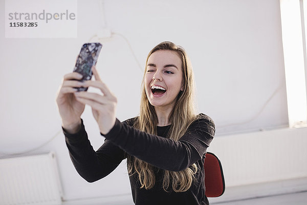 Fröhliche weibliche Bloggerin blinzelt  während sie Selfie durch Smartphone im Büro nimmt.